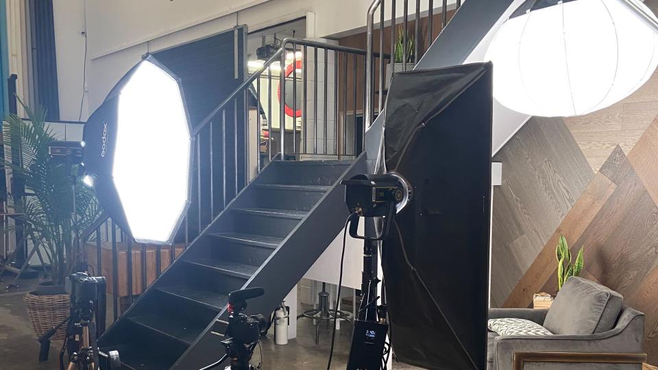 Some of the equipment set up in the shoot in front of a couch; multiple cameras and lighting rigs with cables.