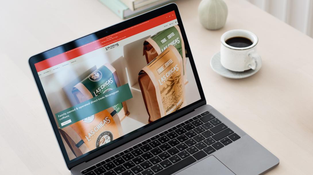 Las Chicas del Café Homepage on a laptop sitting by a coffee mug