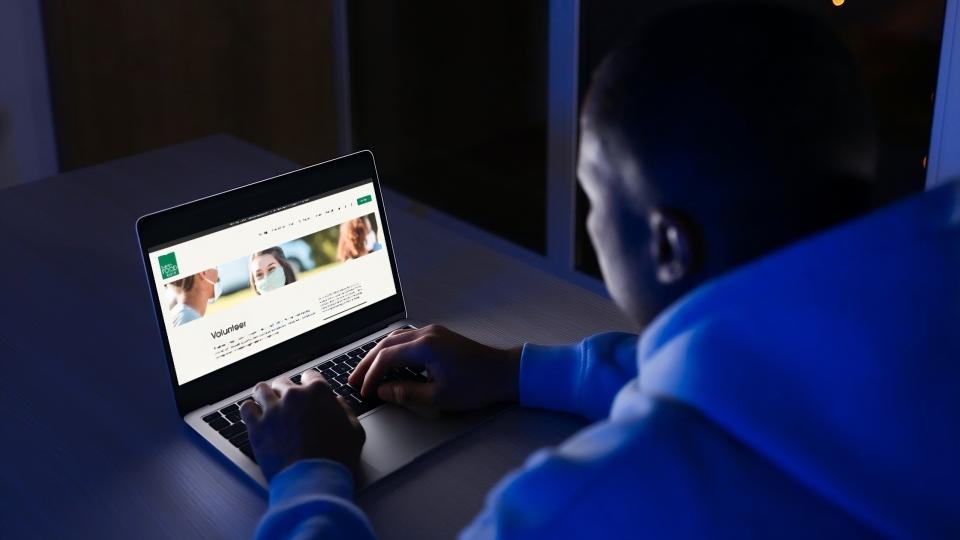 A person in a blue sweater looks at the London Food Bank volunteer page displayed on a laptop.