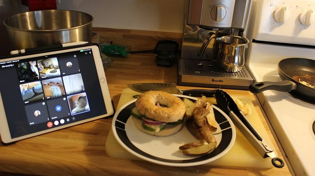 A bagel sandwich on a plate beside an iPad with a zoom call